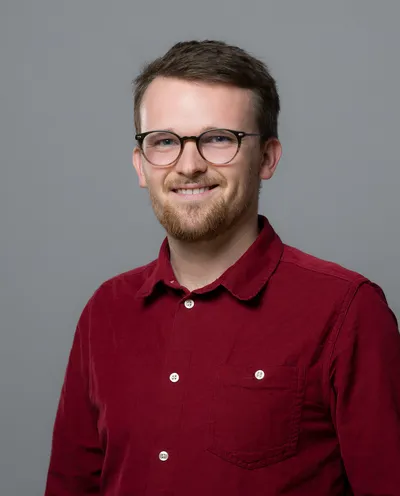 Max Jonas Behrens, bearded and wearing round glasses and a dark red corduroy shirt, in front of a plain gray studio background